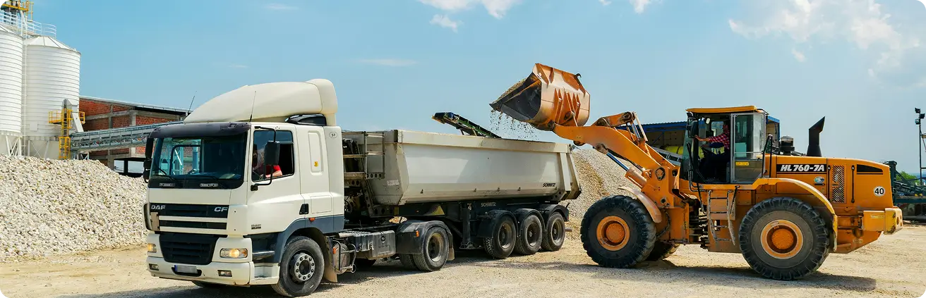 Camião basculante branco a ser carregado por máquina escavadora amarela num estaleiro de construção.